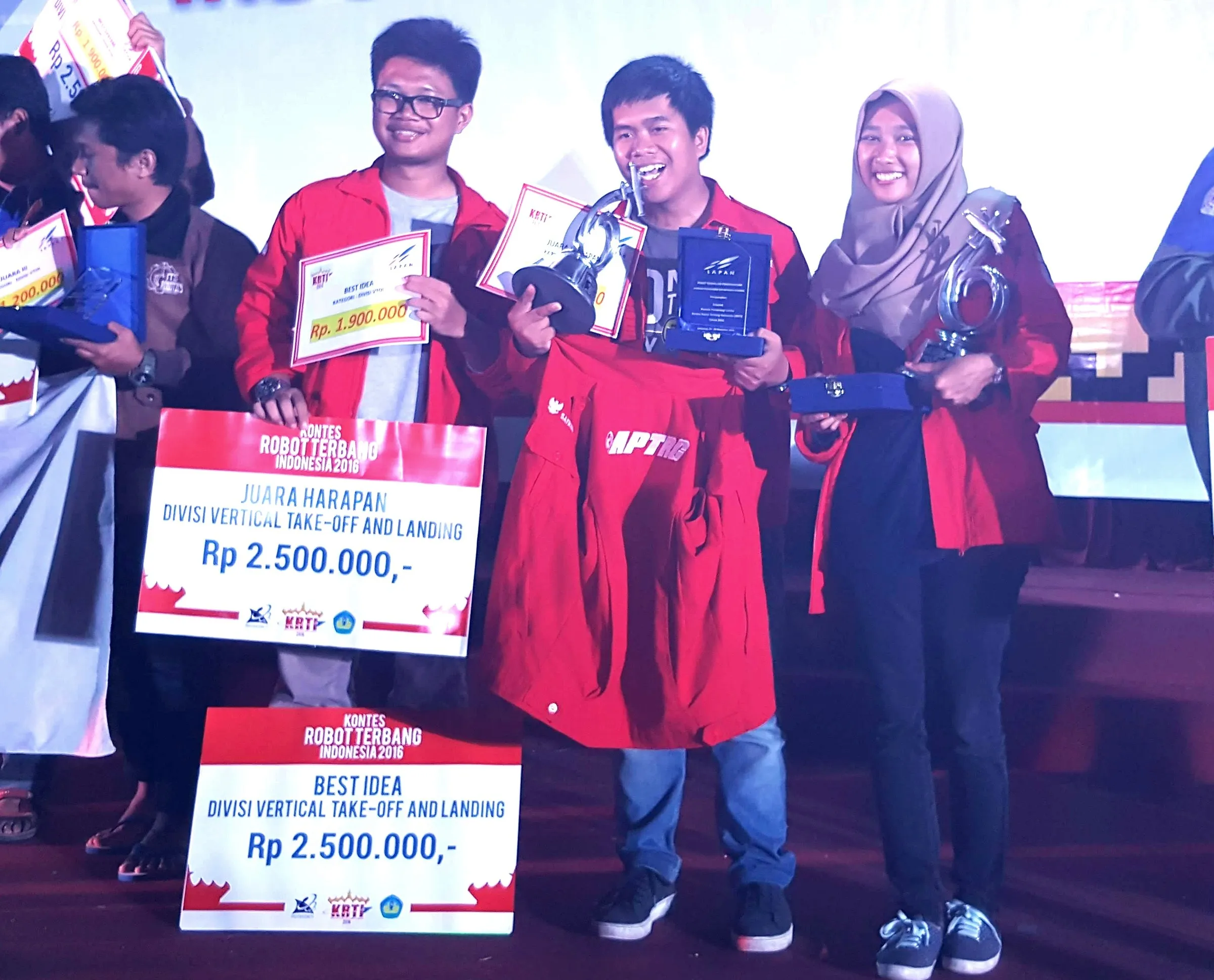 Award ceremony at Kontes Robot Terbang Indonesia 2016. Rasyid, in glasses and a red team jacket, stands with two teammates behind the Best Idea and runner-up boards.