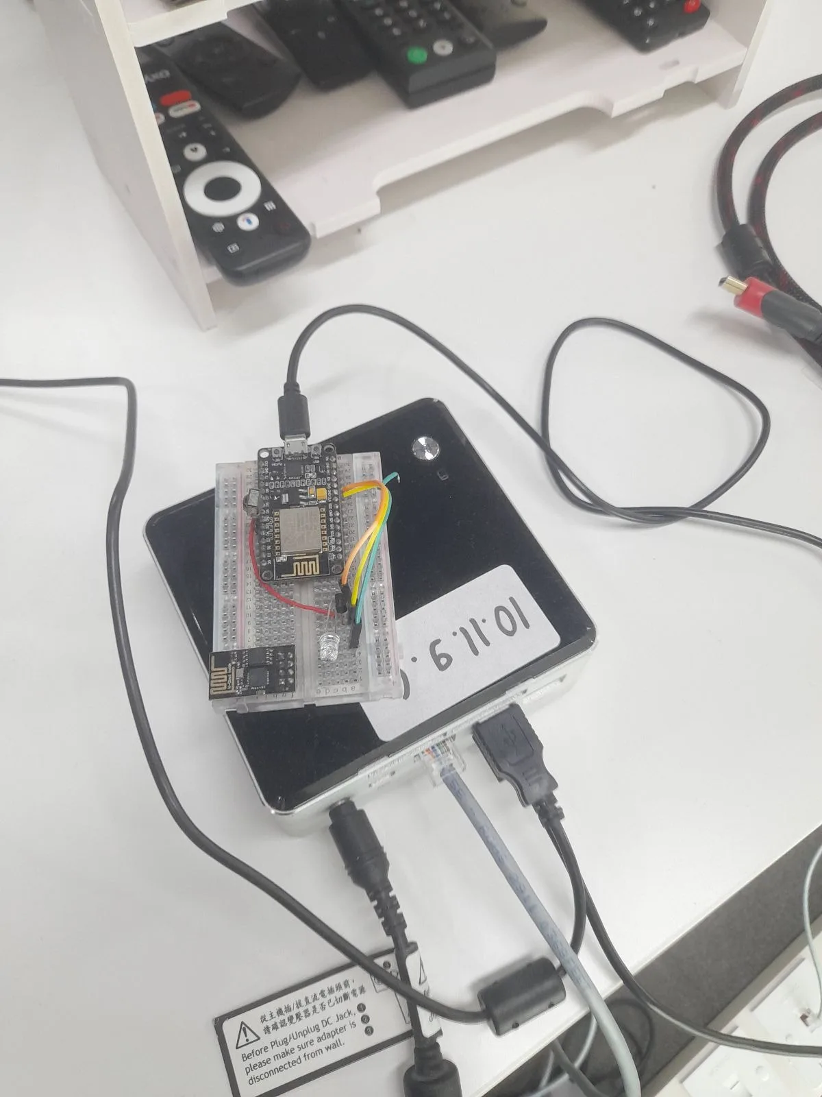 An ESP8266 on a breadboard sitting on top of a small mini PC, with a rack of physical TV remote controls on a shelf in the background.