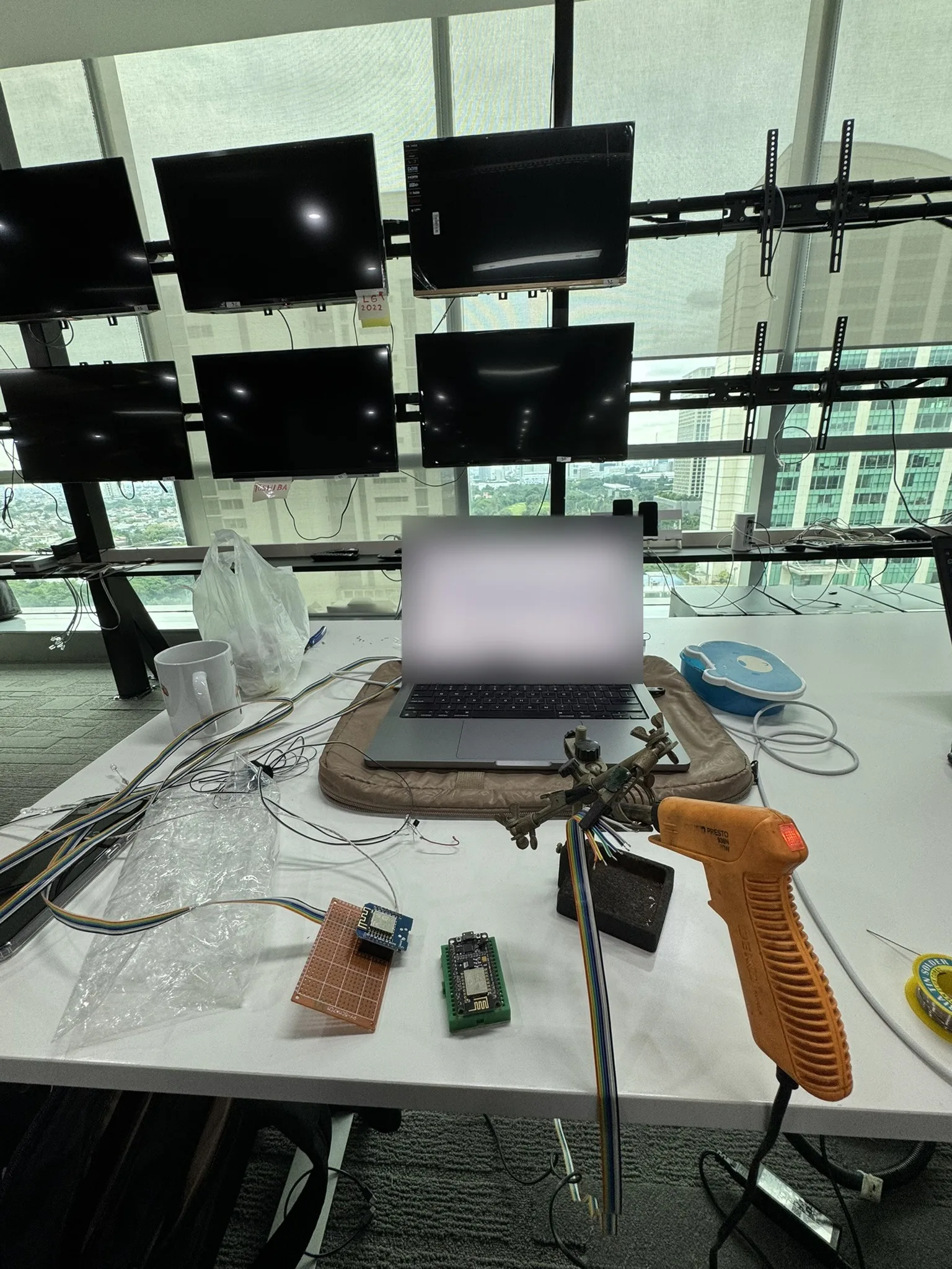 A wall of televisions on custom brackets in front of an office window, with a laptop, ESP8266 boards, ribbon cable and a soldering iron on the desk below.
